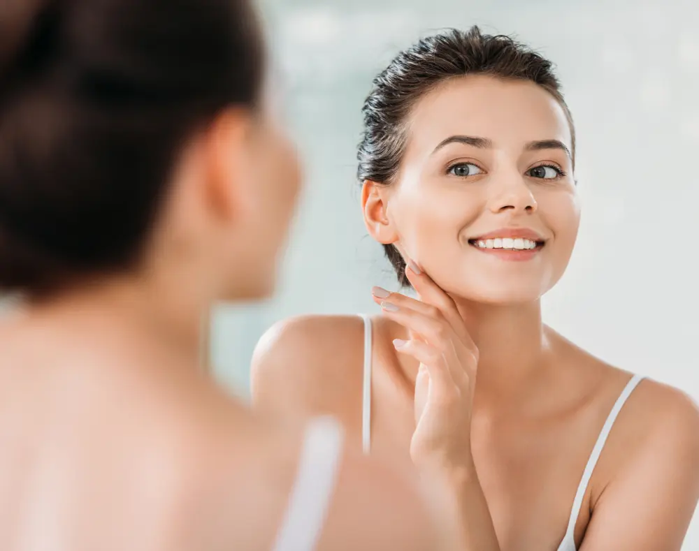 A young woman with short dark hair smiling and touching her face while looking at her reflection in a mirror, with a blurred figure of herself in the foreground.