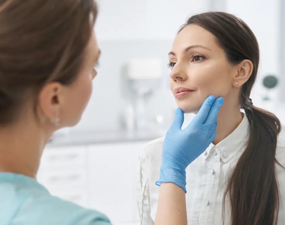 A healthcare professional examines a woman’s face, gently holding her chin with gloved hands in a clinical setting with white background.