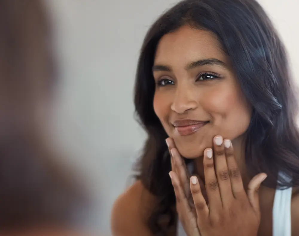 A woman with dark hair gently touches her face with both hands, smiling softly, facing slightly to the right against a neutral background.