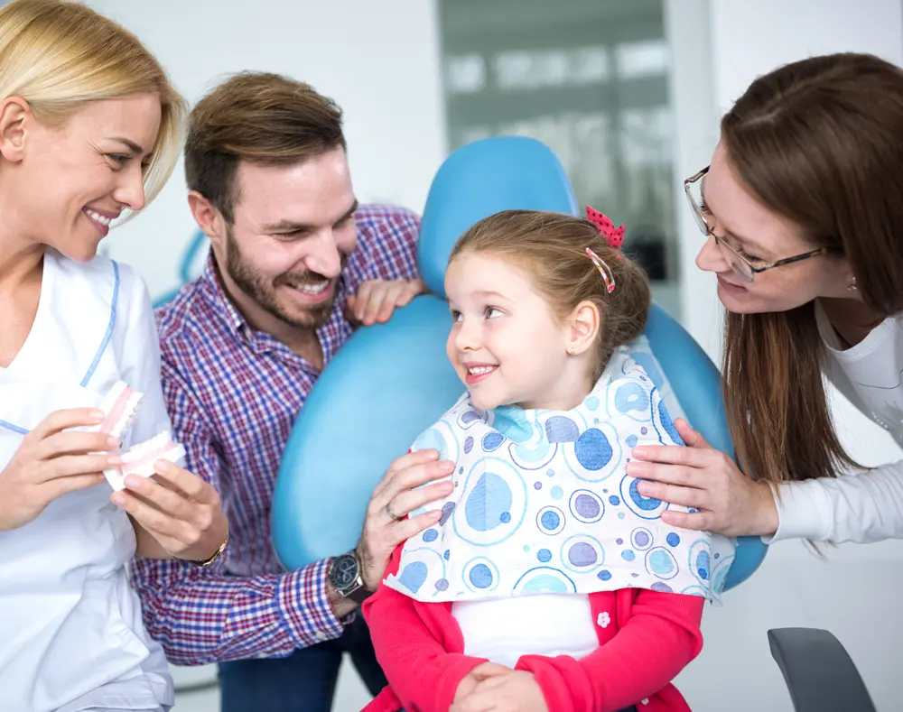 Young girl smiling in dental chair surrounded by two female dental professionals and a male parent, all engaging happily during a dental visit.
