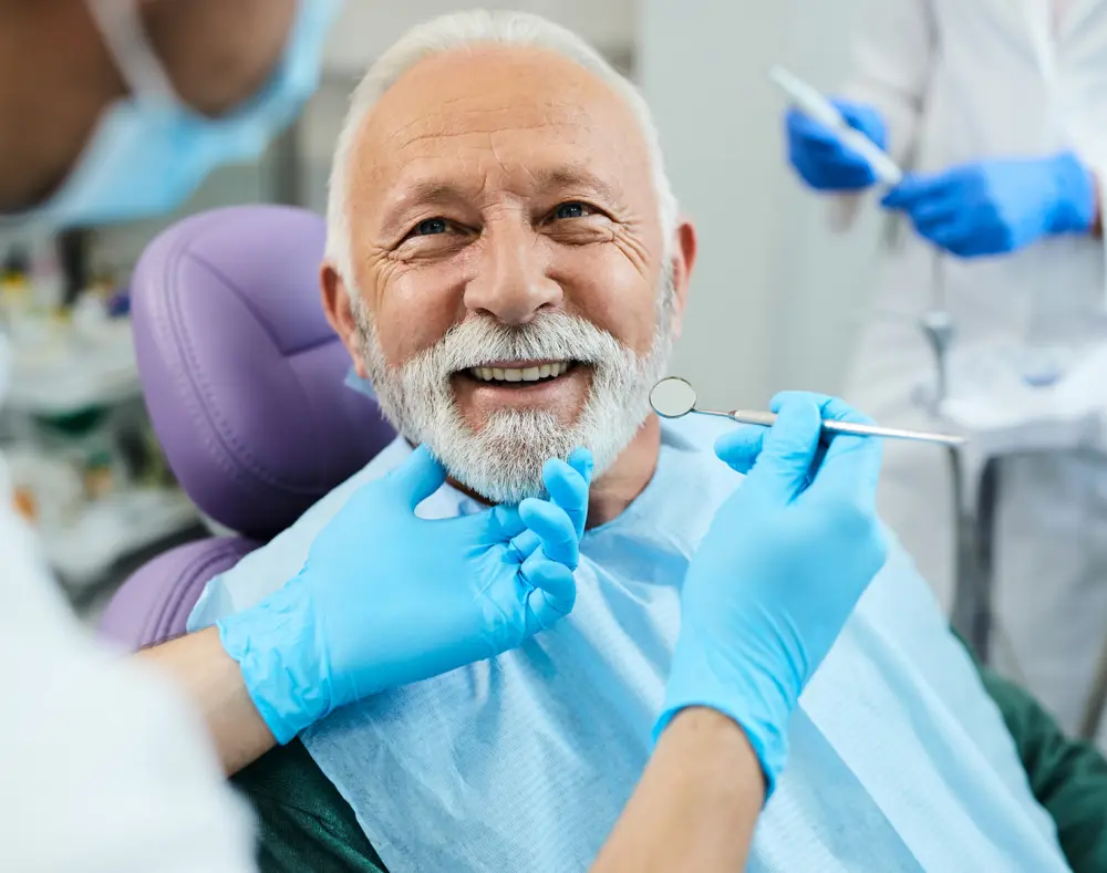 Senior man smiling at the dentist's office, surrounded by dental professionals in protective gear, as the dentist examines his teeth with tools.