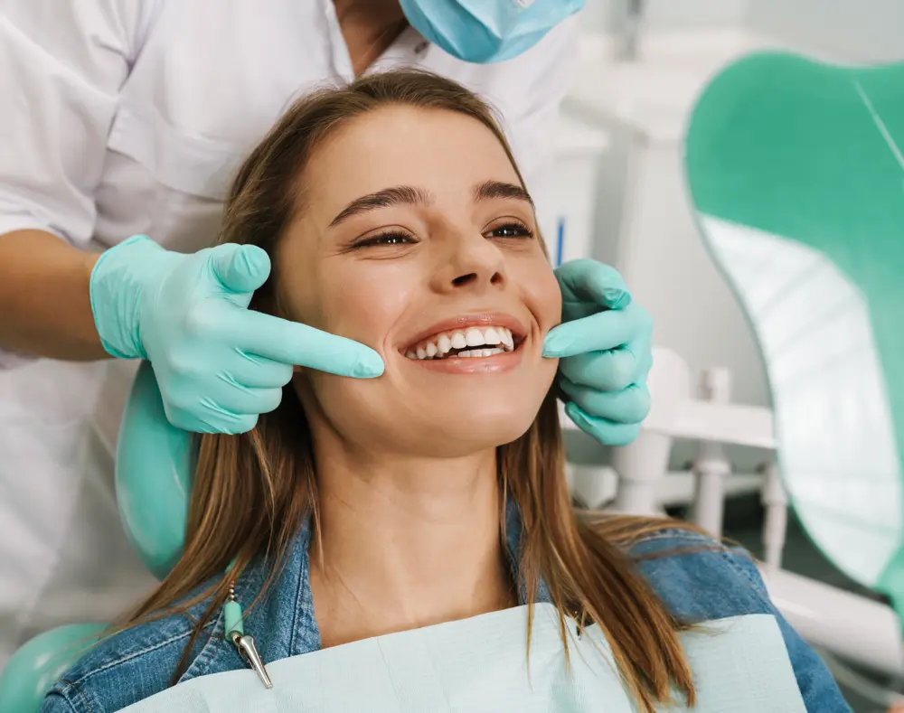 A woman smiling in a dental chair, with a dentist holding her chin and examining her teeth; dental tools and equipment visible in the background.