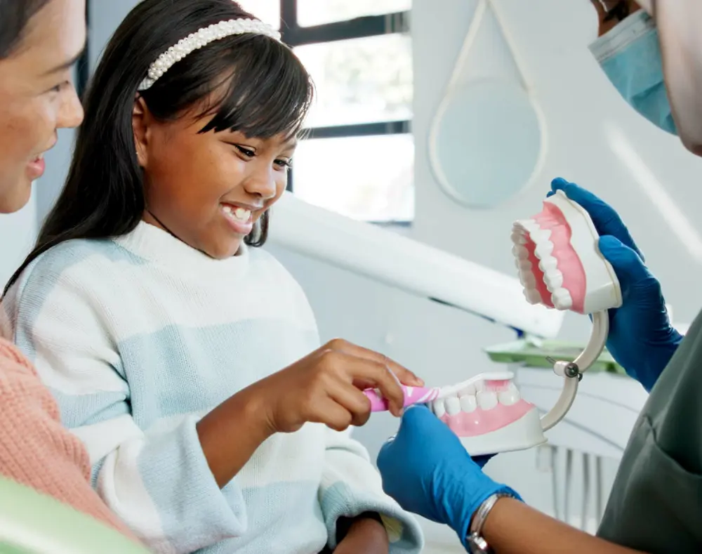 Girl smiling as a dentist demonstrates proper brushing technique with a large dental model, while her mother watches in a bright dental clinic.