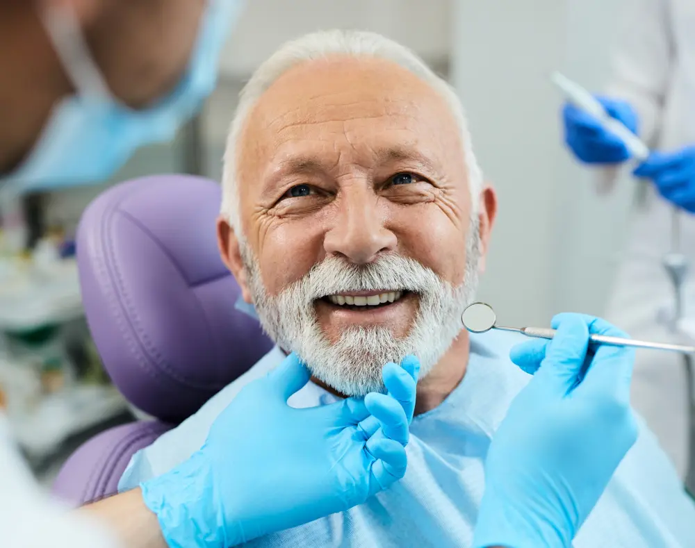 A cheerful elderly man with a white beard at the dentist's, surrounded by dental professionals in blue gloves, with the dentist examining his chin.