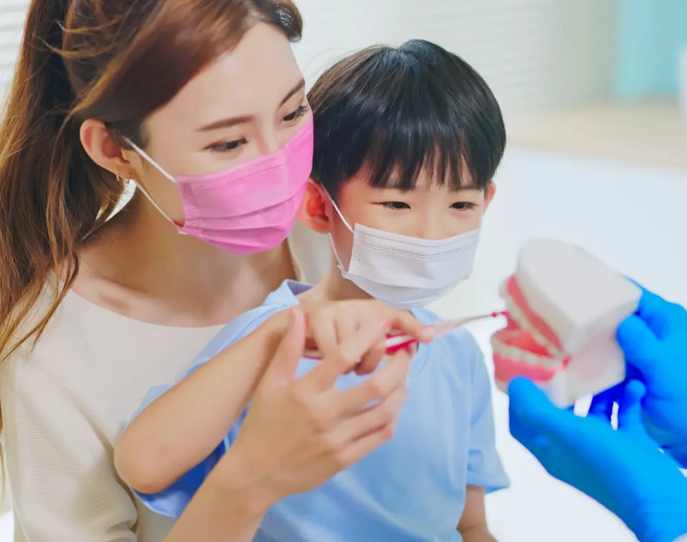 A young boy and woman with masks brushing a large dental model, supervised by a person wearing blue gloves in a brightly lit dental clinic.