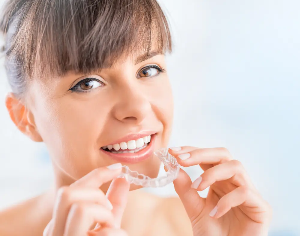 A smiling woman with brown hair holds a clear dental aligner near her teeth against a light background.