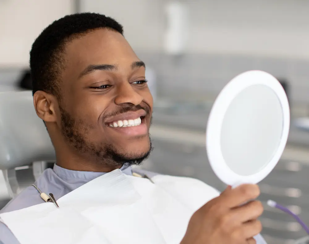 Smiling man in a dental chair holding a mirror, looking at his reflection with a happy expression, in a dental clinic setting.