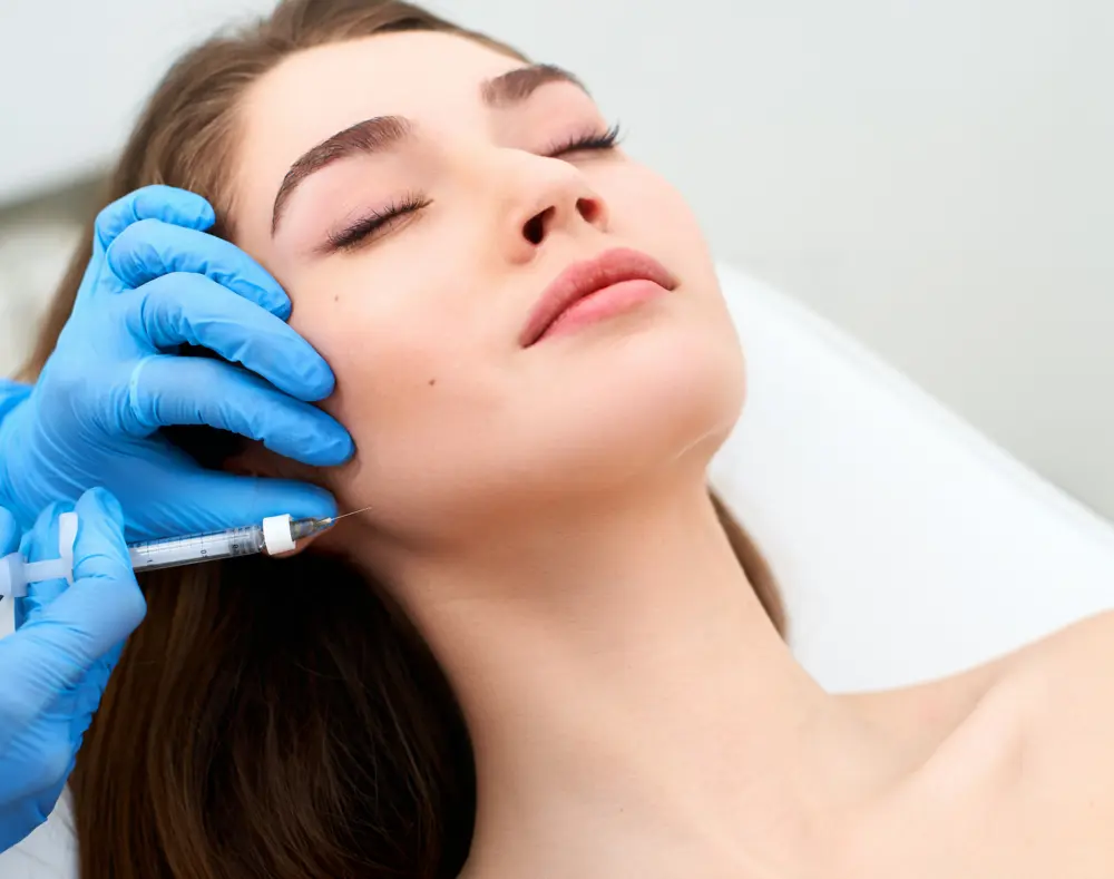 A woman with closed eyes receives a facial treatment from a professional wearing blue gloves, using a syringe near her cheek in a clinical setting.