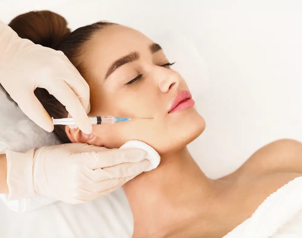 Woman receiving a facial injection while lying down with eyes closed, a medical professional wearing gloves administers the treatment.