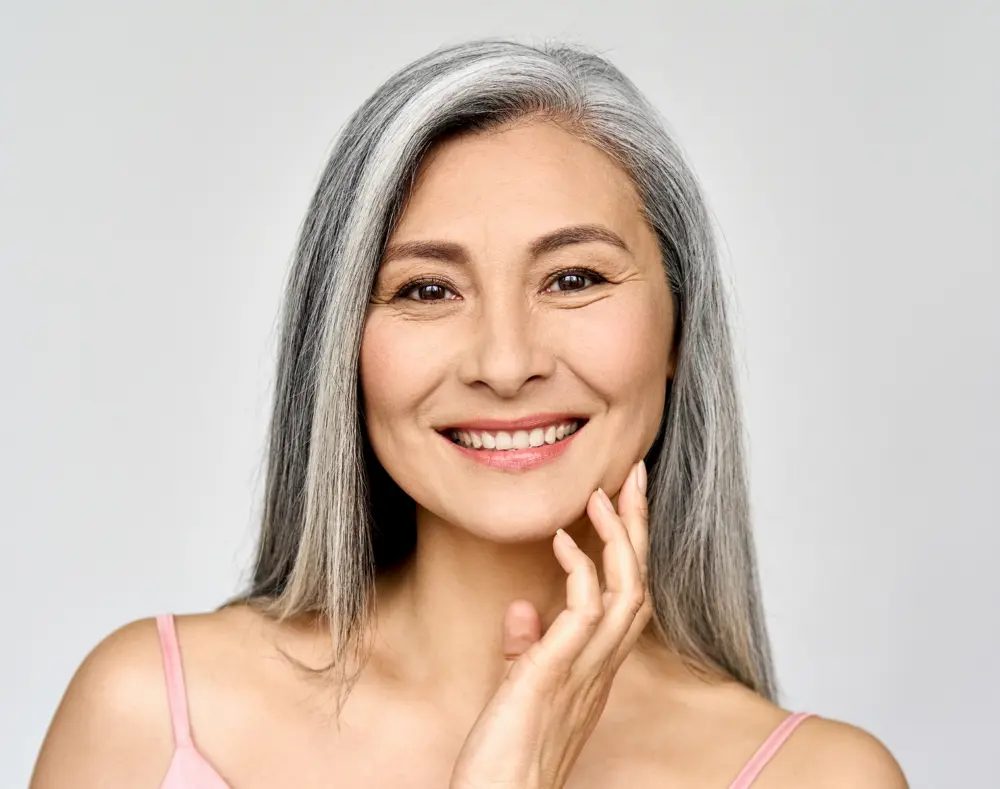 A smiling mature woman with long gray hair, wearing a pink spaghetti strap top, touching her face gently, against a plain light gray background.