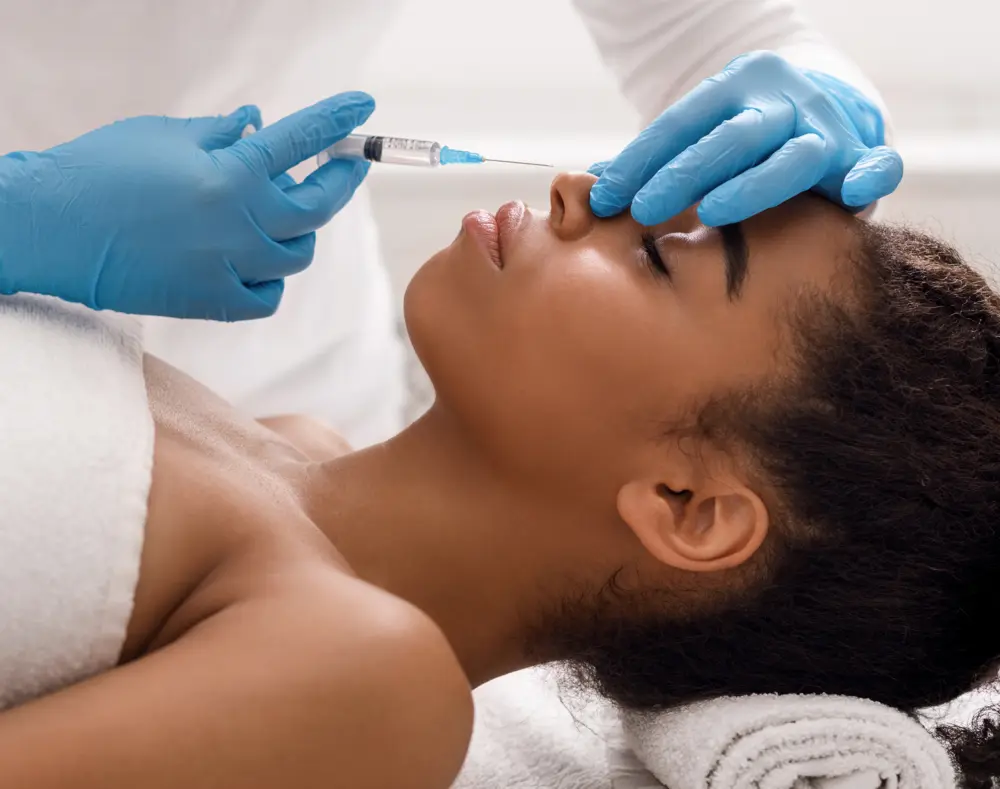 Person receiving a cosmetic injection on her face while lying down, with a healthcare professional wearing blue gloves administering the treatment.