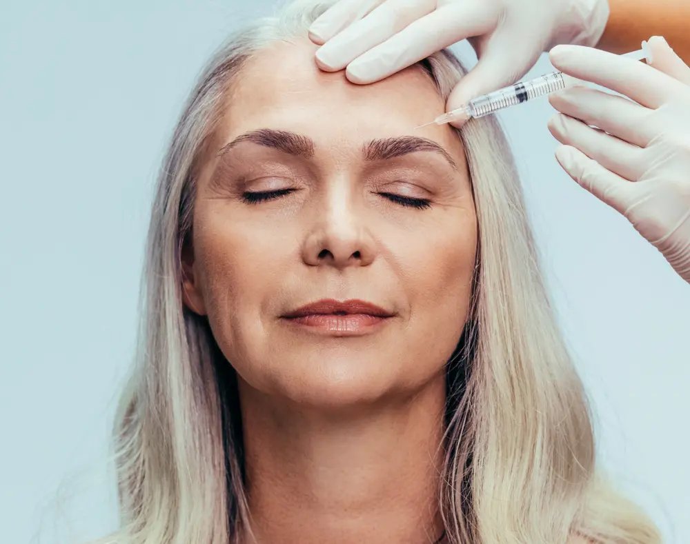 Older woman with closed eyes receives a forehead injection from a healthcare professional wearing gloves.