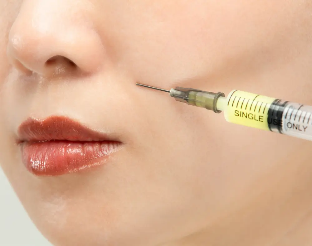 A close-up of a woman's face with a focus on her lips and nose, while a syringe approaches her cheek, suggesting a cosmetic or medical procedure.