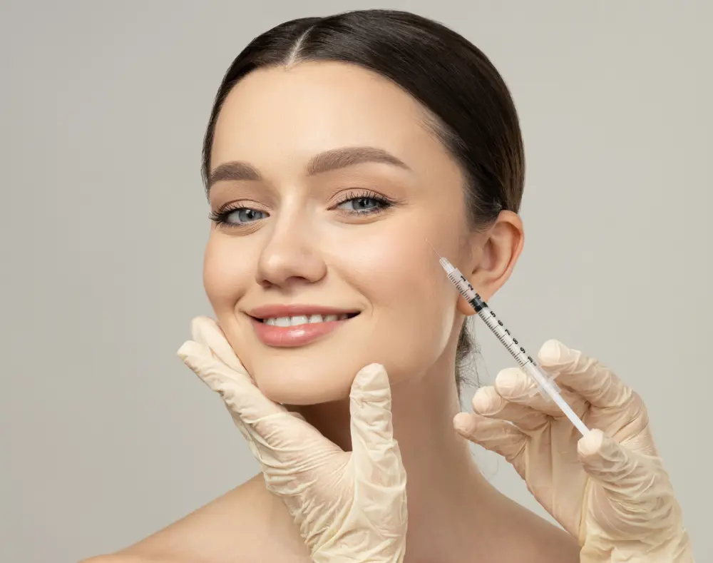 A woman with blue eyes receiving a cosmetic injection in her cheek from gloved hands, smiling gently against a neutral background.