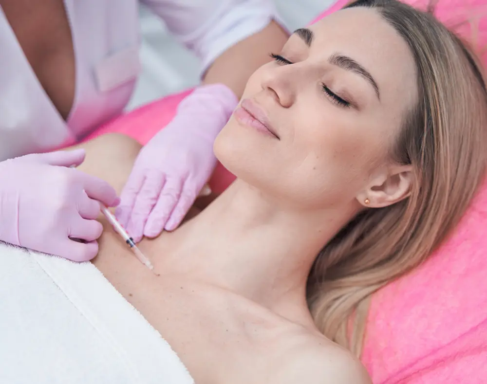 Woman receiving a cosmetic injection in her neck, lying with eyes closed, with a medical professional wearing pink gloves administering the treatment.
