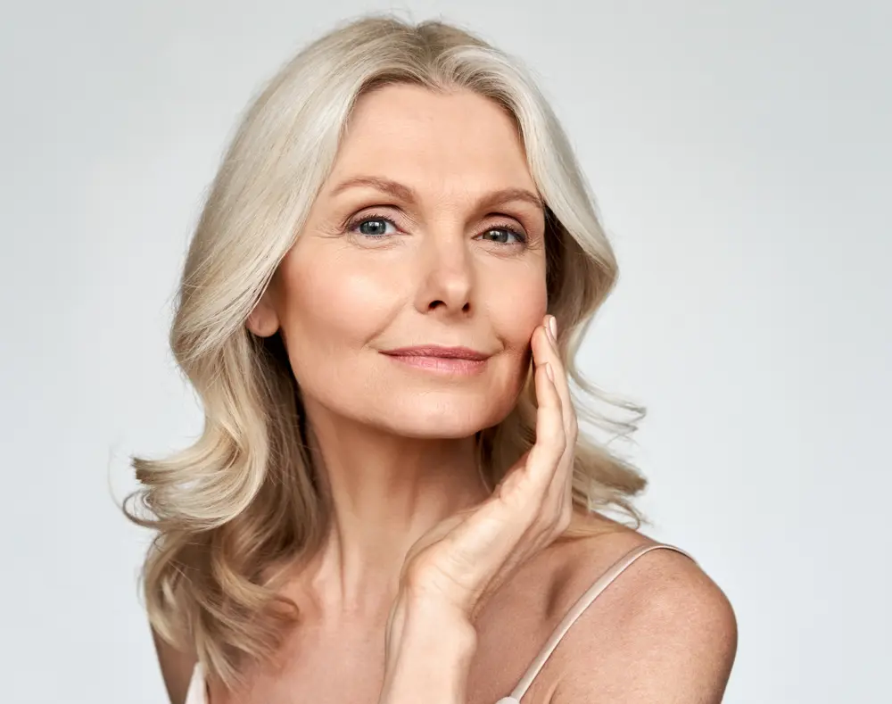 A close-up portrait of a smiling mature woman with long, wavy silver hair, gently touching her face with her right hand against a plain light background.