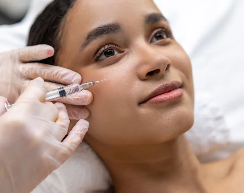 A woman receiving a cosmetic injection to her cheek, lying down with her head tilted slightly, while a gloved hand administers the treatment.