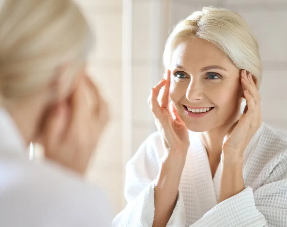 An older woman with platinum blonde hair smiling gently as she looks at herself in a mirror, touching her temples with both hands in a bright, softly lit room.