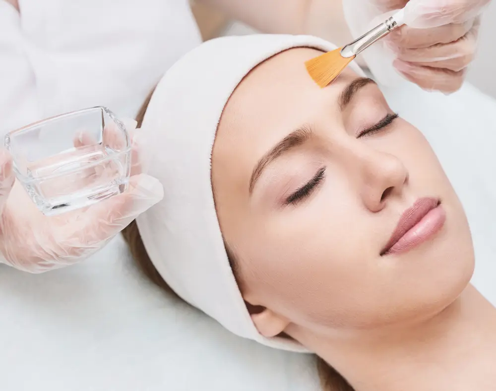 A woman receives a facial treatment, with a skincare professional applying a product using a brush while holding a glass of water. She is relaxed with her eyes closed.