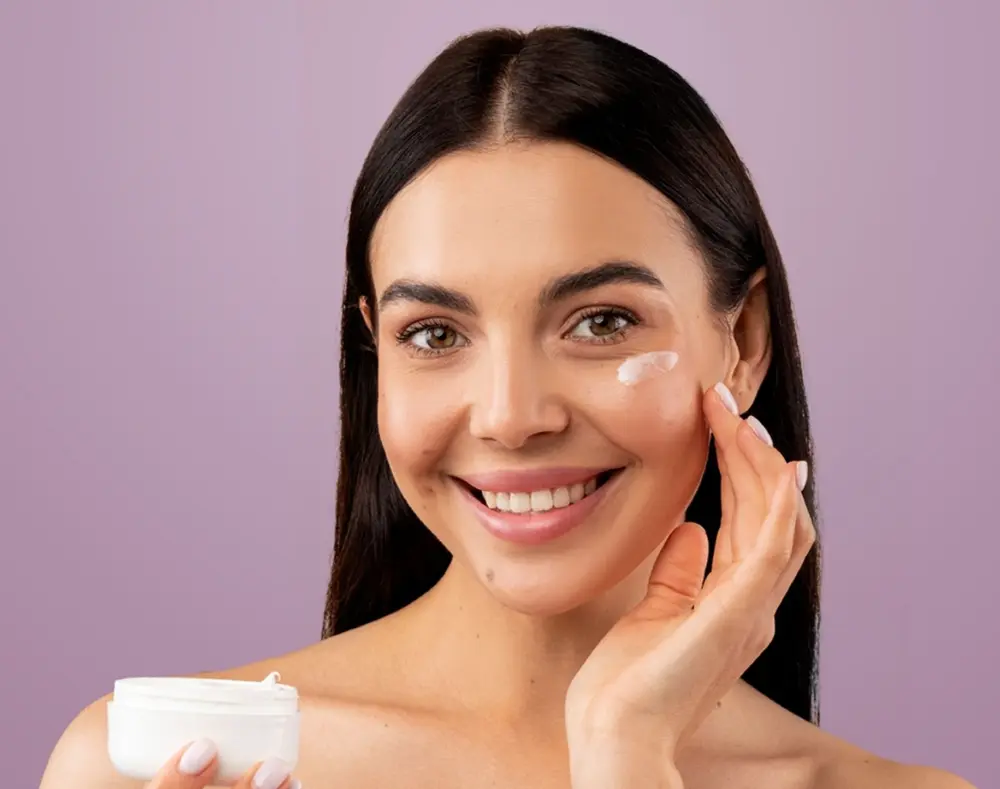 Woman smiling while applying facial cream on her cheek against a purple background.