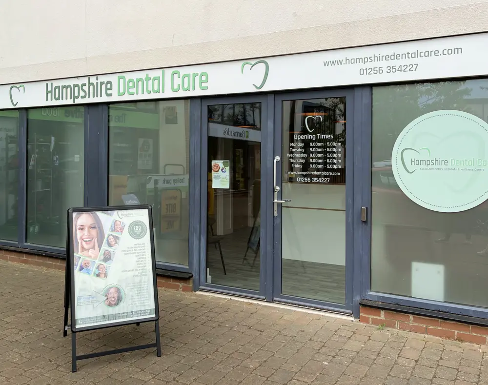 Exterior view of Hampshire Dental Care clinic with glass doors, signs displaying hours, and a promotional stand outside.