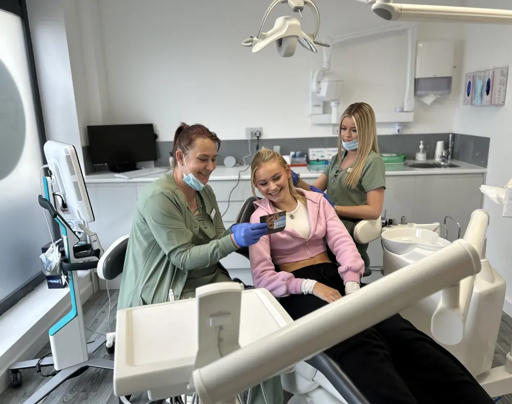 A teenage girl in a pink hoodie sitting in a dental chair, smiling while looking at a photo, with two dental professionals in scrubs and masks assisting.
