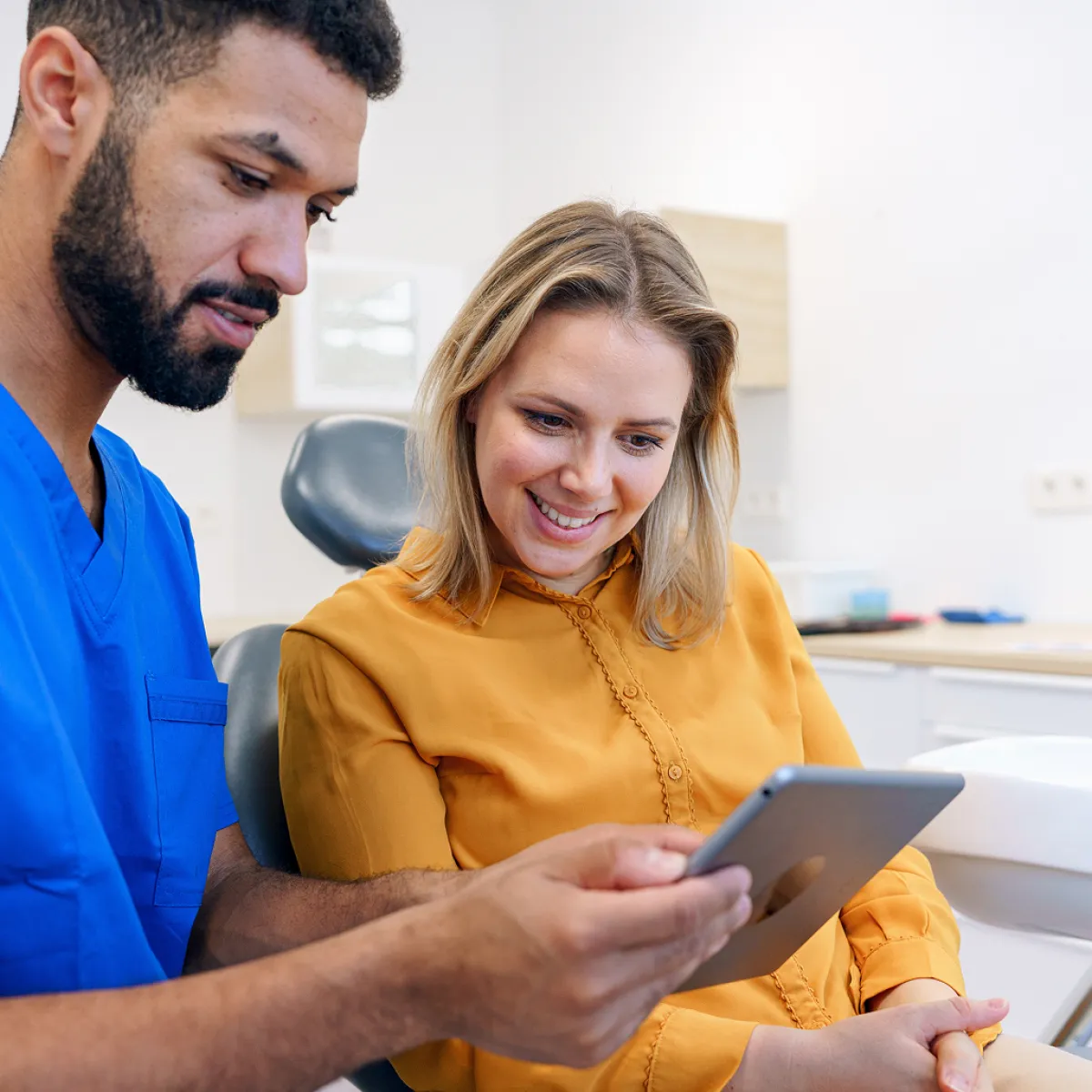 A healthcare professional in blue scrubs and a woman in an orange blouse look at a tablet together in a clinical setting, smiling.