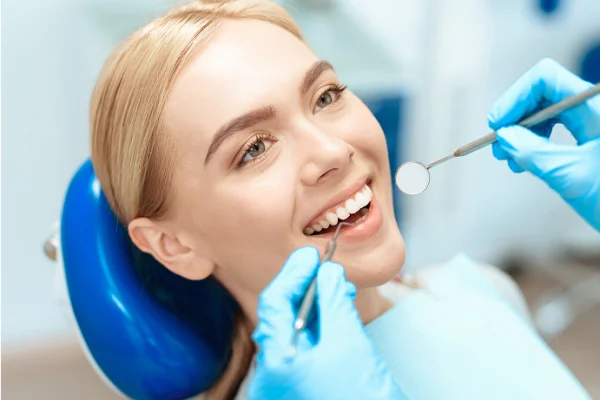 Woman at dentist's office receiving dental check-up, lying in a dental chair, smiling while a dentist examines her teeth with dental tools.