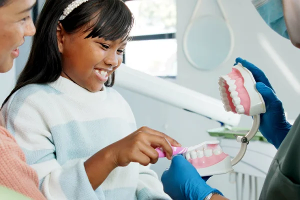 Girl smiling as a dentist demonstrates proper brushing technique with a large dental model, while her mother watches in a bright dental clinic.