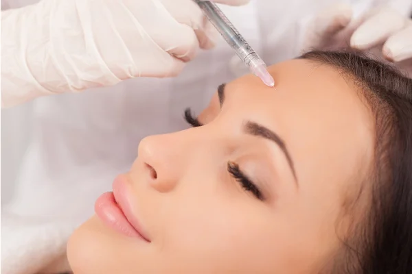 Woman receiving a facial injection with a syringe while relaxing with eyes closed. The practitioner wears gloves and white attire.