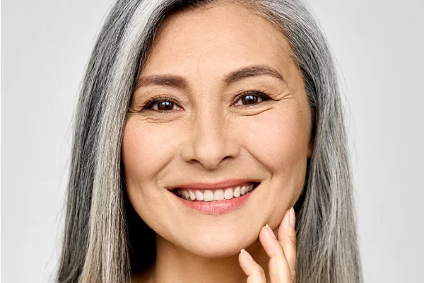 A close-up of a smiling mature woman with long gray hair, touching her face gently, against a plain light background.