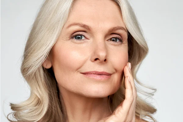 A mature woman with long, wavy gray hair gently touches her face, smiling softly against a plain light background.