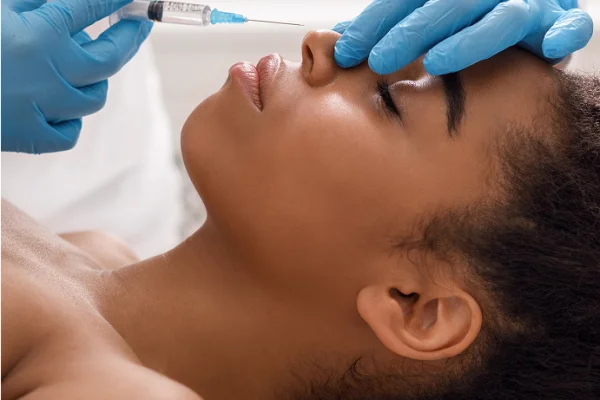 A woman receiving a cosmetic injection in her forehead from a medical professional wearing blue gloves.