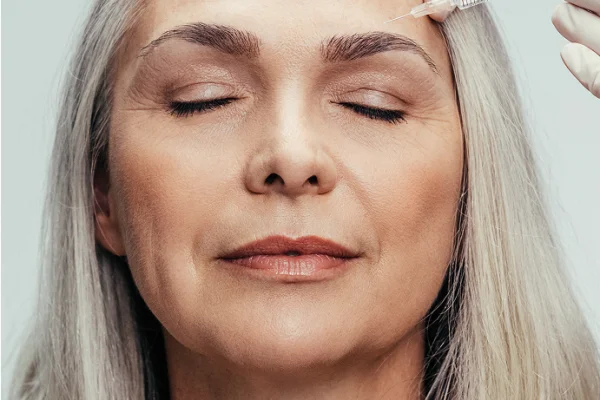 A mature woman with closed eyes receiving a cosmetic injection on her forehead as a healthcare professional administers the treatment with a syringe.