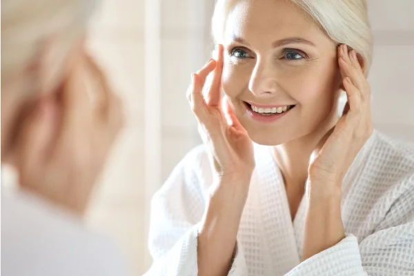 A smiling woman with blonde hair adjusting her hair, wearing a white robe, sitting in front of a mirror in a bright, softly lit room.