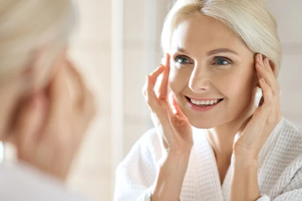 A woman with blonde hair smiling at herself in a mirror while touching her face, in a bright, well-lit room.