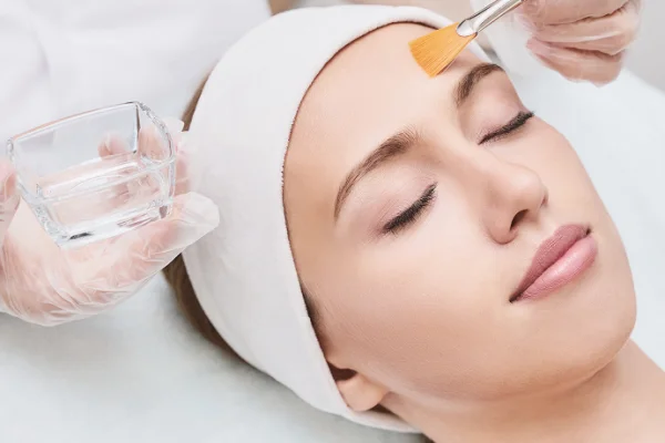 A woman receives a facial treatment, lying peacefully with a headband, while a skincare professional applies a product with a brush.