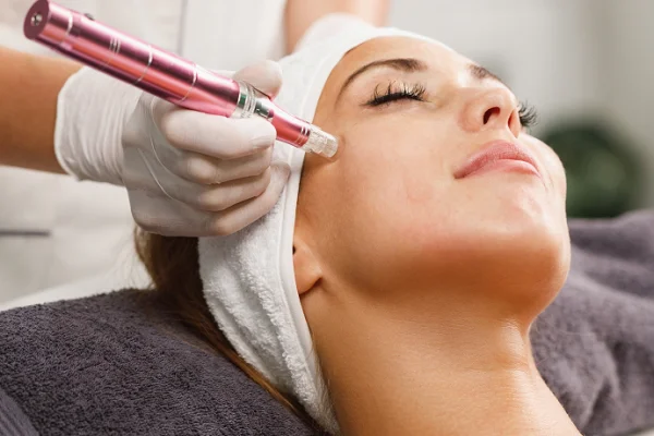 Woman receiving facial treatment with a pink device applied to her cheek, lying down with a towel wrapped around her head, eyes closed in a spa setting.