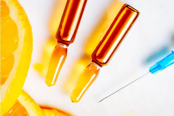 Two amber glass ampoules and a blue syringe needle on a white surface, with orange slices in the foreground.