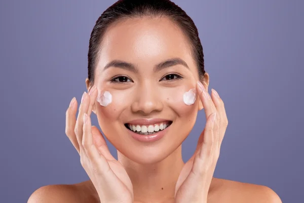 A smiling young woman with dark hair, applying cream to her cheeks against a purple background.