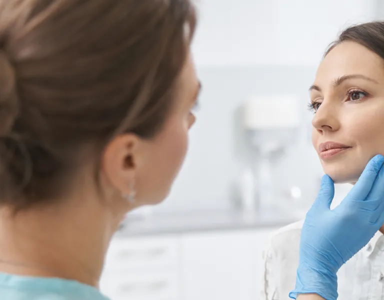 Medical professional examining a woman's face in a clinical setting, with the doctor gently touching the woman's chin and cheek.