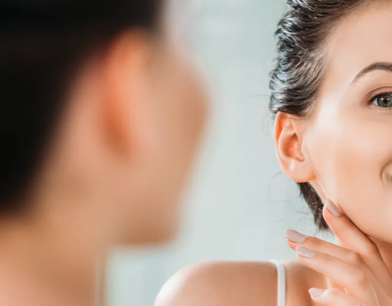 A woman smiling while touching her chin, looking at her reflection in a mirror, with a clear focus on her face and expression.
