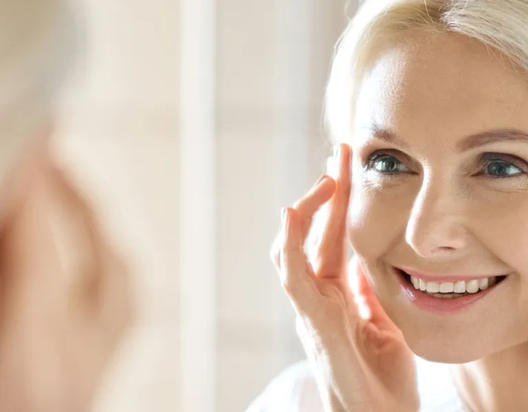 A woman with fair skin and blonde hair looks at herself in a mirror, gently touching her face with both hands and smiling.