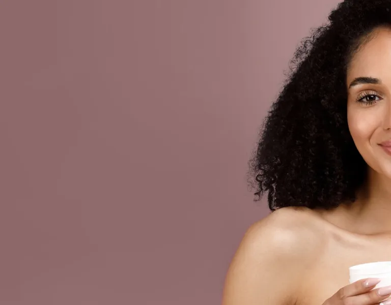 A woman with curly hair smiling and holding a jar of skincare cream against a soft pink background.