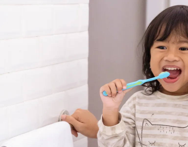 A smiling woman and young girl brushing their teeth together in a bathroom, both holding toothbrushes, with a white tiled wall in the background.