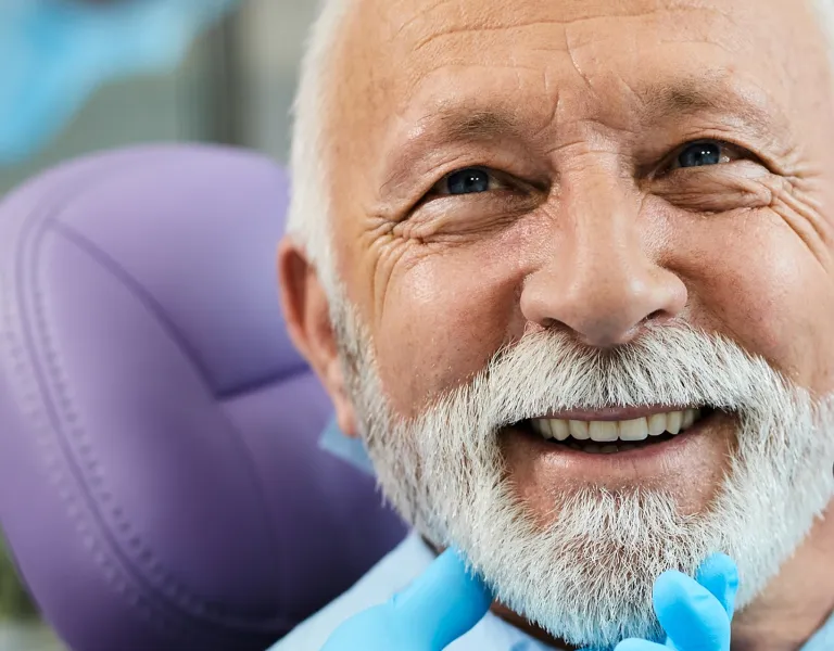 An elderly man with a white beard smiling at the dentist, who is holding a dental mirror near his mouth in a bright dental clinic.