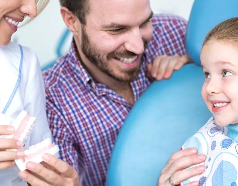 A girl at the dentist's office smiling while holding a dental model, with her parents and a dentist presenting her with a smile.